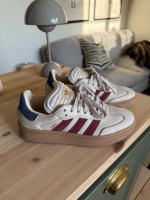 Adidas sneakers med vinröda ränder - Snygga Adidas sneakers i beige läder med klassiska vinröda ränder på sidorna och blå detalj vid hälen. Skorna har gummisula och guldfärgade Adidas-loggor på plösen och sidan. Perfekta för dig som gillar retrostil och streetwear. Använda fåtal gånger på grund av försmå. Utöver det super fina och sköna!