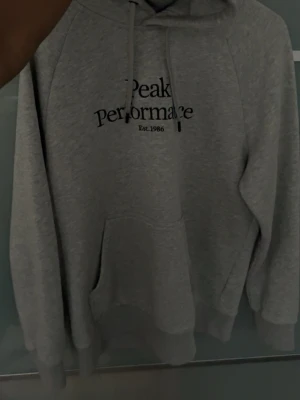 Grå hoodie från Peak Performance - Snygg grå hoodie från Peak Performance med svart broderad logga och text 'Est. 1986' på bröstet. Storlek M men passar S också. Klassisk känguruficka framtill, justerbar huva med dragsko och mjukt material som känns skönt mot huden. Perfekt för en avslappnad och sportig stil. Pris kan diskuteras. 