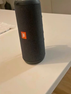 JBL flip 4  - Snygg svart bärbar Bluetooth-högtalare från JBL med cylinderformad design och robust tygmaterial runt om. Utrustad med tydliga knappar för volym och uppspelning samt en praktisk bärrem. Perfekt för att ta med sig överallt och spela musik trådlöst.