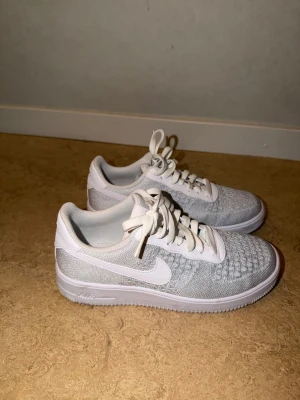Nike Air Force 1 Flyknit vita sneakers - Snygga Nike Air Force 1 Flyknit sneakers i vitt med klassisk låg siluett. Ovandelen är i stickat material som ger en lätt och luftig känsla, med vita skosnören och den ikoniska swoosh-loggan på sidan. Perfekta för dig som gillar stilrena och bekväma sneakers.