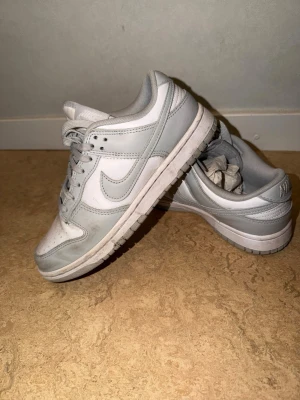 Nike Dunk Low vit/grå sneakers - Nike Dunk Low sneakers i vitt och ljusgrått läder. Klassisk siluett med perforerad tåbox, snörning och ikoniska swoosh-loggan på sidan. Yttersula i gummi för bra grepp. Perfekta för dig som gillar stilrena och tidlösa sneakers.
