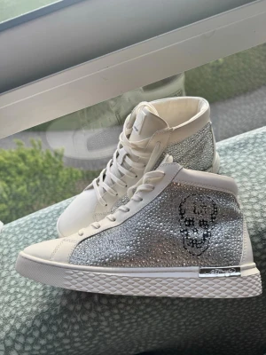 Glittriga vita sneakers från ed hardy - Helt nytt, Snygga vita sneakers från ed hardy med högt skaft och massor av glittrande stenar på sidorna. Skorna har en cool döskalle i svart på sidan och vita snören. Perfekta för dig som vill sticka ut med en lyxig och unik stil.