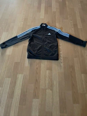 Svart Adidas träningsjacka - Säljer en svart träningsjacka från Adidas med klassiska vita ränder längs ärmarna och dragkedja framtill. Jackan har hög krage och är tillverkad i ett glansigt syntetmaterial som passar perfekt till träning eller sportiga outfits.