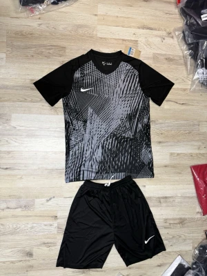 Svart Nike träningsset herr - Snyggt svart träningsset från Nike med t-shirt och shorts. T-shirten har ett grafiskt mönster i grått och vitt samt vit Nike-logga på bröstet. Shortsen är helsvarta med vit logga och har dragsko i midjan. Perfekt för fotboll eller gym.