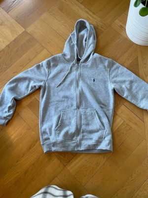Grå zip hoodie från Polo Ralph Lauren - Klassisk grå hoodie med dragkedja från Polo Ralph Lauren. Hoodien har huva med dragsko, två stora fickor fram och den ikoniska lilla loggan broderad på bröstet. Tillverkad i mjuk bomullsblandning som är perfekt för chill dagar.