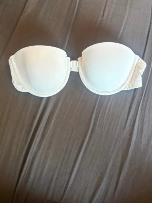 Vit axelbandslös bh - Säljer en vit/ljusbeige axelbandslös bh med bygel och vadderade kupor. Bh:n har en smidig knäppning fram och  kupor som ger ett snyggt lyft. Perfekt under toppar och klänningar med bara axlar. Materialet känns mjukt och stretchigt. Endast testad