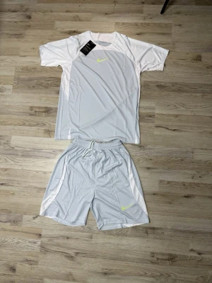 Nike ljusgrått träningsset herr - Snyggt ljusgrått träningsset från Nike med t-shirt och shorts. Setet har vita detaljer på sidorna och neongult Nike-logga på både tröjan och shortsen. Tillverkat i ett lätt och snabbtorkande funktionsmaterial som passar perfekt till träning.