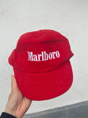 Röd Marlboro keps  - Rare röd keps från Marlboro i mjuk manchester med vit broderad logga framtill. Klassisk böjd skärm och justerbar passform bak. Ikonisk! 
