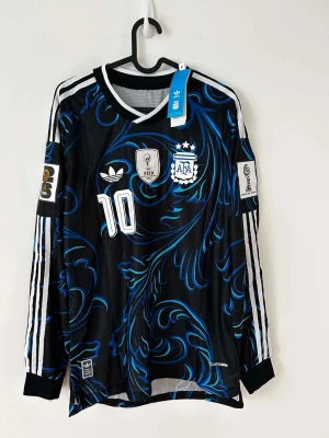 Argentina långärmad Messi tröja spelar version! - Snygg långärmad fotbollströja från Adidas för Argentina, Messi #10. Svart bas med blått och vitt mönster, officiella FIFA och AFA-emblem samt vita ränder på ärmarna. Tillverkad i lätt och ventilerande material, perfekt för träning eller match. Spelar version;