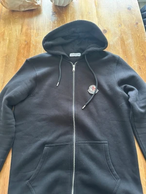 Svart Moncler hoodie med dragkedja - Säljer en svart hoodie från Moncler med dragkedja framtill och klassisk Moncler-logga på bröstet. Hoodien har huva med dragsko och två stora fickor framtill. Tillverkad i mjukt bomullsmaterial som känns riktigt skönt mot huden.