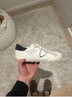 Philippe Model vita sneakers med blå detalj - Snygga vita sneakers från Philippe Model med blå häl och grå mockadetaljer. Skorna har en klassisk låg siluett, vita snören och den ikoniska sköldloggan på sidan. Perfekta för dig som gillar stilrena och trendiga sneakers.