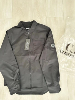 Svart overshirt från C.P. Company - Stilren svart overshirt från C.P. Company med dragkedja framtill, bröstficka med lock och ikonisk linsdetalj på ärmen. Overshirten har klassisk krage och knappdetaljer vid ärmslut. Perfekt för lager-på-lager och streetwear.