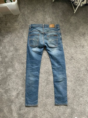 Blå slim fit jeans från Nudie Jeans - W30 L32. Säljer ett par blå slim fit jeans från Nudie Jeans med klassisk femficksmodell och snygga slitningar på framsidan. Jeansen har normal midja, smala ben och är tillverkade i mjuk denim. Perfekta för en avslappnad och trendig look.