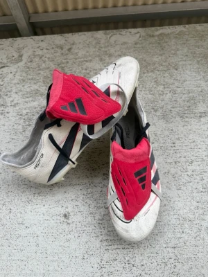 Adidas Predator vita fotbollsskor - Säljer ett par klassiska Adidas Predator fotbollsskor i vitt med svarta och röda detaljer. Skorna har en ikonisk röd plös med svarta Adidas-ränder. Använda men i superfint skick. 