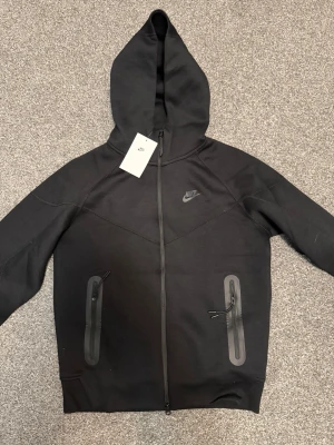 Svart Nike Tech Fleece hoodie - Nike Tech Fleece hoodie i svart med hel dragkedja och två fickor med dragkedja framtill. Snygg minimalistisk design med Nike-logga på bröstet och huva. Tillverkad i mjukt och lätt material som passar perfekt till streetwear-stilen.