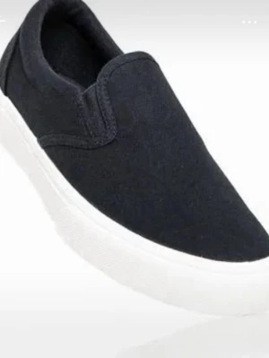 Svarta slip-on sneakers - Stilrena svarta slip-on sneakers med vit sula. Ovandelen är i textilmaterial och skon har en enkel, minimalistisk design utan snörning. Perfekta för dig som gillar en clean look och vill ha något lätt att ta på sig.