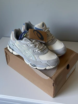 Asics vita och grå sneakers - Snygga sneakers från Asics i vitt och ljusgrått med meshpaneler och detaljer i syntet. Klassisk Asics-logga på sidan och bekväm, vadderad insida. Skorna har en chunky sula och sportig look, perfekta för en trendig streetstyle.