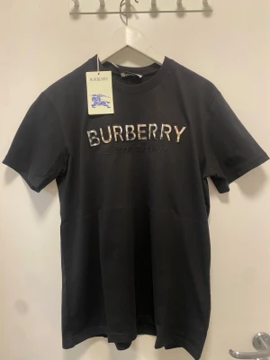 Svart Burberry t-shirt med logga - Snygg svart t-shirt från Burberry med klassisk logga broderad i vitt och grått på bröstet. T-shirten har rund halsringning och korta ärmar. Tillverkad i mjuk bomull för en bekväm känsla. Perfekt för dig som vill ha en stilren och exklusiv look. Passar perfekt till sommaren och till nästan alla outfits. Helt ny med prislapp. Kom med ett prisförslag om du är sugen, eller köp direkt såklart 😉 skickar samma dag!