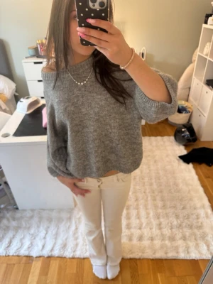 Grå off shoulder stickad tröja - Mysig grå stickad tröja med off shoulder-design och bred halsringning. Tröjan har en avslappnad passform och vida ärmar som ger en trendig look. Perfekt att matcha med jeans eller kjol för en chill stil.