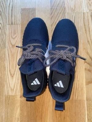 Svarta Adidas sneakers med mesh - Snygga svarta sneakers från Adidas med ovandel i mesh och klassiska tre ränder på sidan. Skorna har svart sula med ribbad struktur och svartvita mönstrade skosnören. Perfekta för en sportig och trendig look.