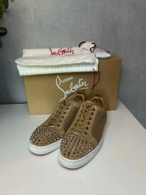 Beige sneakers från Christian Louboutin - Snygga beige sneakers från Christian Louboutin med vita sulor och coola silverfärgade nitar på tån. Skorna har ett texturerat material i mocka och läderlook, samt matchande beige skosnören. Perfekt för dig som vill sticka ut med din stil. 40 passar 41