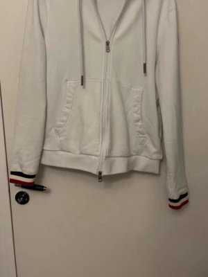 Moncler Hoodie  - Säljer en vit hoodie från Moncler med dragkedja framtill, huva med dragsko och fickor på magen. 