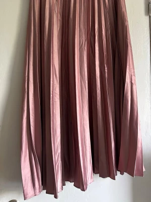 Lång plisserad rosa kjol från Zara - Säljer en lång plisserad kjol från Zara i en snygg rosa nyans. Kjolen har ett glansigt material och vackra veck som ger ett elegant fall. Finns skavank på ett ställe (bild finns)