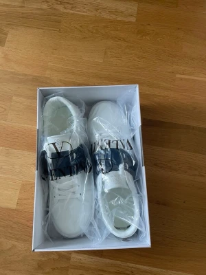 Valentino Open - Snygga vita sneakers från Valentino med bred mörkblå rem över vristen och klassisk snörning. Skorna har en stilren och modern design med logga på plösen. Tillverkade i slätt skinn för en exklusiv känsla.