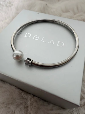Silverfärgat armband från Edblad - Snyggt armband i silverfärg från Edblad med en stor vit pärla och en gnistrande klar sten i ändarna. Stilren och modern design som passar perfekt till din outfit. Armbandet är i metall och har en öppen form som gör det lätt att ta på. ❗️(kan sänka priset)❗️ boxen följer med!!