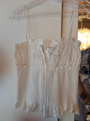 Vit linnetopp med volang och knyt - Söt vit linnetopp med tunna axelband, volangkant nedtill och smockad struktur. Toppen har en liten knytning framtill som ger en extra detalj. Perfekt för varma dagar och enkel att styla till jeans eller kjol.