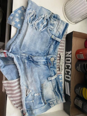 Ljusblå slitna jeansshorts - Säljer ett par ljusblå jeansshorts  med slitna detaljer och råa kanter. Shortsen har hög midja och en cool patch med stjärnor och ränder på bakfickan. Perfekta för sommaren och ger en avslappnad streetstyle-look.