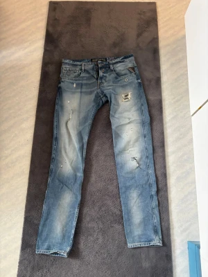 Blå slitna jeans med raka ben - Säljer ett par blå Replay anbass jeans med slitningar och trasiga detaljer på framsidan. Byxorn är lite halv skum men ändå lite avslappnad, så inte för tajta. Storlek 30 i midja och 32 i längd 