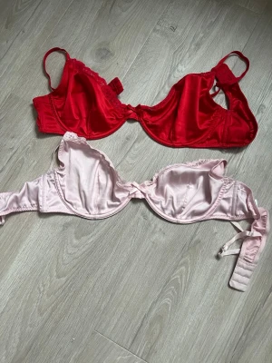 Två bhar i satin, röd och ljusrosa - Säljer två bhar i glansigt satinmaterial, en i rött och en i ljusrosa. Båda har bygel och justerbara axelband. Den röda har spetsdetaljer vid kuporna och den rosa har en liten rosett framtill. Perfekta för dig som gillar färg och mjuka material.