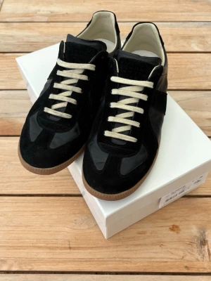 Svarta sneakers från Maison Margiela - Svarta sneakers från Maison Margiela med klassisk låg siluett. Skorna har en mix av mocka och slätt läder, vita snören och beige yttersula i gummi. Insidan är fodrad i vitt läder för extra komfort. Perfekta för en clean och stilren look.