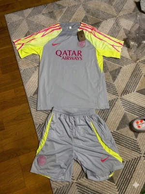 PSG träningsset grå/gul Nike - Säljer ett PSG träningsset från Nike i grått med neon-gula detaljer och rosa loggor. Tröjan har korta ärmar med rosa ränder och Qatar Airways-tryck på bröstet. Matchande shorts med PSG-logga och Nike swoosh. Perfekt för fotbollsträning.