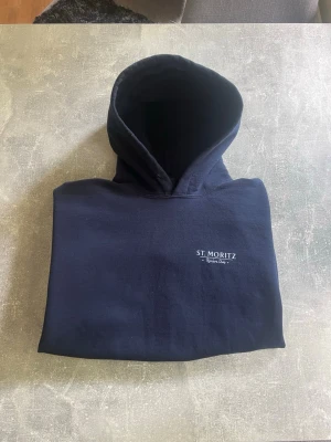 Mörkblå hoodie Reviere club - Snygg mörkblå hoodie från Reviere club med texten St. Moritz på framsidan och en bild på baksidan. Perfekt för vardagen men kan användas till nästan allt.