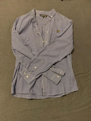 Blårutig skjorta från Lyle & Scott - Snygg blårutig skjorta från Lyle & Scott med klassisk button-down krage och gul logga på bröstet. Skjortan har vita knappar och är långärmad. Perfekt för dig som gillar stilrena och tidlösa plagg.
