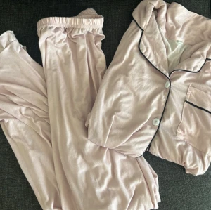 Ljusrosa pyjamas set - Mysig ljusrosa pyjamas i mjukt material med långärmad skjorta och byxor. Skjortan har vita knappar, svart passpoal längs krage och ficka samt en bröstficka. Perfekt för chillkvällar hemma.