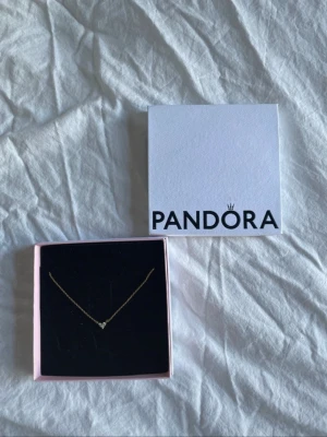 Guldhalsband med hjärta från Pandora - Snyggt och stilrent halsband i guld från Pandora med ett litet hjärta dekorerat med gnistrande stenar. Kedjan är tunn och elegant, perfekt för att lyfta vilken outfit som helst. Kommer i originalask från Pandora. Nypris är 1500 och den är använd cirka en vecka.