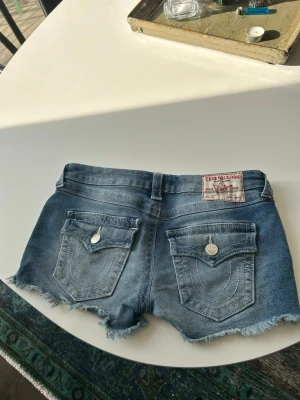Blå jeansshorts från True Religion joey cut of w 27 - Säljer ett par blå jeansshorts från True Religion med råa, fransiga kanter och klassiska bakfickor med lock och knapp. Mycket bra skick nypris 1099 kr. Perfekta för sommaren!