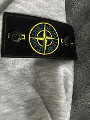 Grå hoodie från Stone Island - Snygg grå hoodie från Stone Island med den klassiska svarta patchen och loggan på ärmen. Hoodien har dragsko i huvan och är tillverkad i mjukt bomullsmaterial. Perfekt för dig som vill ha en stilren och trendig look.