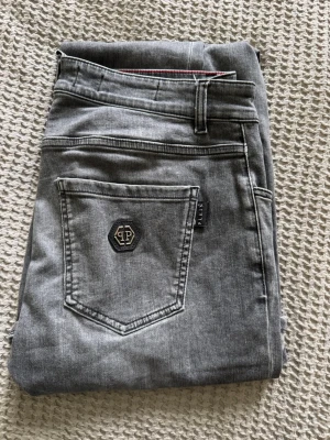Feta grå jeans från Philipp Plein - Snygga grå jeans från Philipp Plein med klassisk femficksmodell och coolt logomärke på bakfickan. Jeansen har raka ben och detaljer som metallknappar och små färgade nitar vid fickan. Riktigt bra skick och kvitto + taggar finns. Nypris är runt 3000