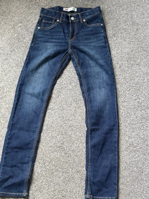 Levi's blå raka jeans - Säljer ett par klassiska blå jeans från Levi's med rak passform och fem fickor. Jeansen har snygga kontrastsömmar och en ikonisk läderpatch bak i midjan. Tillverkade i slitstark denim som passar perfekt till en avslappnad stil. Jeansen är aldrig använda och där med helt nya.