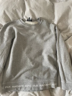 Grå sweatshirt från A.P.C. - Snygg grå sweatshirt från A.P.C. med rund halsringning och långa ärmar. Diskret logga och texten 'RUE MADAME A.P.C.' längs nederkanten. Tillverkad i mjukt bomullsmaterial som känns skönt mot huden. Perfekt för en clean och stilren look.
