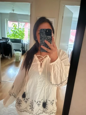 Vit blus med broderi och snörning - Bohemisk vit blus med svarta broderade detaljer längs ärmar och nederkant. Blusen har långa ärmar, snörning med tofsar vid halsen och en lös, flowy passform. Perfekt för dig som gillar en avslappnad och trendig stil.