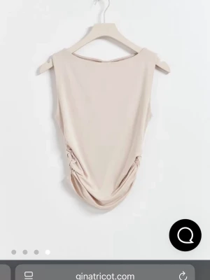 Beige ärmlös topp från Gina Tricot - Snygg beige ärmlös topp från Gina Tricot med draperad effekt och rynkade sidor. Toppen har en rund halsringning och croppad passform, perfekt att styla med höga jeans eller kjol. Tillverkad i mjukt stretchigt material.