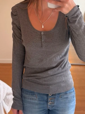 Grå långärmad tröja med knappar - Mysig grå långärmad tröja med rundad urringning och fyra dekorativa knappar framtill. Tröjan har ribbade muddar vid ärmslut och nederkant, samt lätt puffade axlar för en trendig look. Perfekt att matcha med jeans.