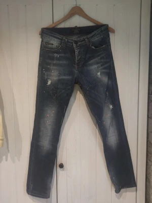 Philipp Plein Couture blå jeans - Säljer ett par blå jeans från Philipp Plein Couture med coola slitningar och färgstänk för en edgy look. Jeansen har klassisk femficksmodell, röd kontrastsöm i midjan och raka ben. Perfekta för dig som vill sticka ut med din stil.