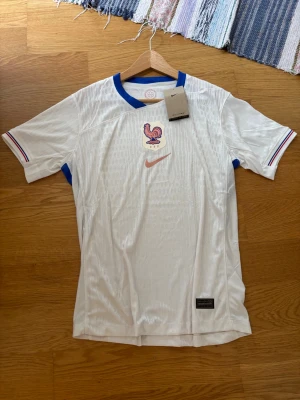 Vit Frankrike fotbollströja Nike - Snygg vit fotbollströja från Nike med Frankrikes landslagsemblem och blå detaljer vid kragen och ärmsluten. Tröjan har korta ärmar och är tillverkad i ett lätt, ventilerande material som passar perfekt till träning eller match. Helt ny!!