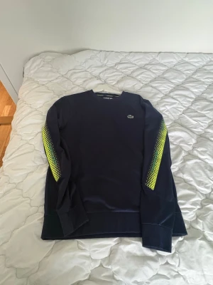 Svart sweatshirt från Lacoste - Cool svart sweatshirt från Lacoste med neongula detaljer längs ärmarna och klassisk krokodil-logga på bröstet. Tröjan har rund halsringning och ribbade muddar vid ärmslut och nederkant. Perfekt för en sportig och trendig look. Storleken passar väldigt bra för S me